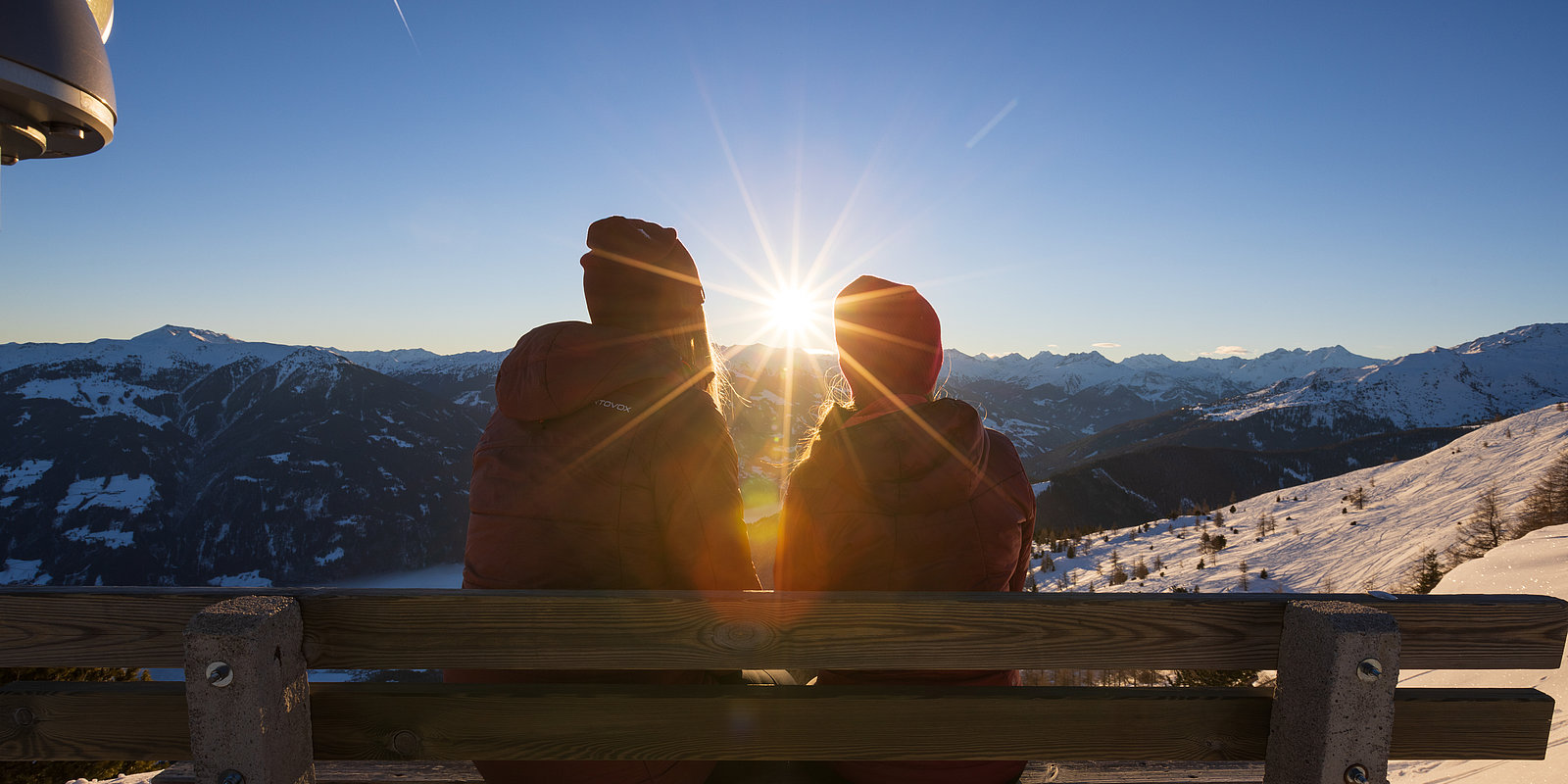 Zwei junge Frauen beim Sonnenaufgang, sitzend auf der Bank beim Gipfelkreuz