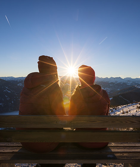 Zwei junge Frauen beim Sonnenaufgang, sitzend auf der Bank beim Gipfelkreuz